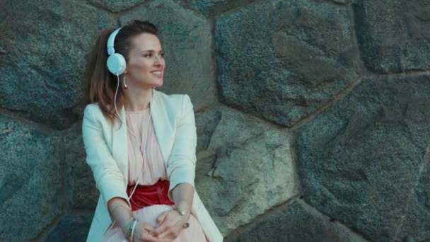 ピンクのドレスを着た流行の女性と街の白いジャケットを笑顔石壁に対するヘッドフォンで音楽を聞いて — ストック動画