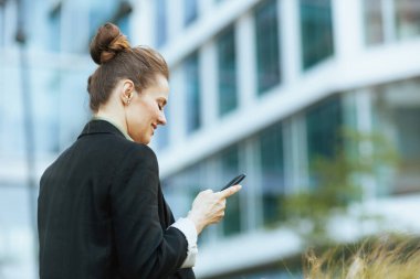 Ofis binasının yanındaki modern iş kadınının arkasında siyah ceketli akıllı telefon kullanarak görülmüş..