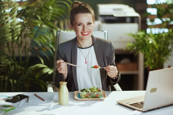 持続可能な職場 幸せな現代的な中年会計士女性でグレービジネススーツで現代緑のオフィスでラップトップ食べてサラダ — ストック写真