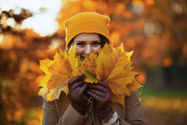 城市公园里穿着米黄色外套 头戴橙色帽子 头戴秋天黄色叶子的快乐女性 — 图库照片