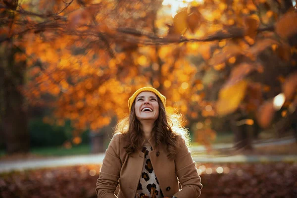 十一月 穿着米黄色外套 头戴橙色帽子 面带微笑的中年女性在城外的城市公园里抛撒秋天的树叶 — 图库照片