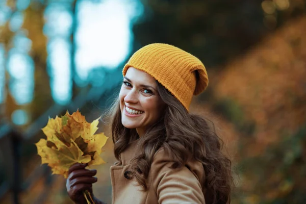 十一月 快乐的40岁女人 穿着米黄色的外套 头戴橙色的帽子 秋天的黄叶飘扬在城外的城市公园里 — 图库照片