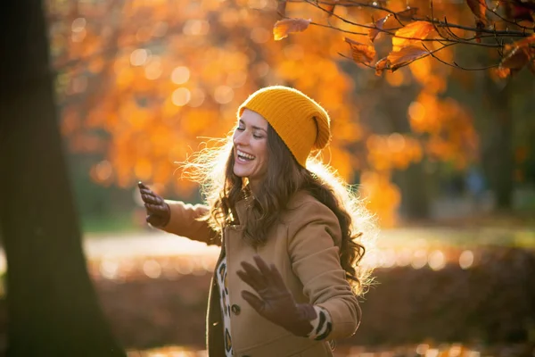 十一月 穿着棕色外套 头戴黄色帽子 面带微笑的女人在城市公园外面跳舞 — 图库照片