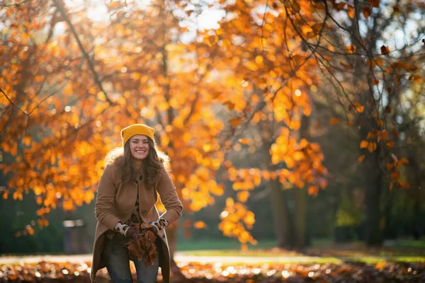 穿着米黄色外套 头戴橙色帽子的优雅的女性在城市公园的室外抛撒秋天的树叶 — 图库照片