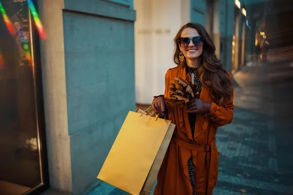 城市里 穿着棕色风衣 头戴购物袋 头戴秋天黄色叶子的快乐时尚女性正在商店附近徘徊 — 图库照片