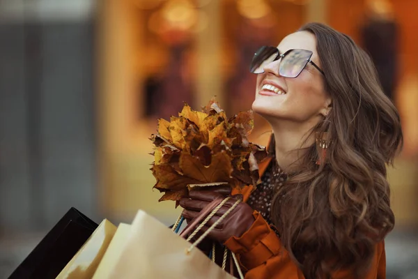城市里 穿着橙色风衣 头戴购物袋 头戴秋天黄色叶子的优雅而快乐的女人正在商店附近徘徊 — 图库照片