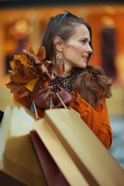 こんにちは ショッピングバッグと秋の黄色い葉が街の店の近くにある茶色のトレンチコートの若い女性 — ストック写真