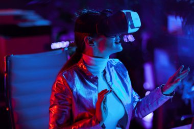 Neon metaevren fütüristik konsepti. Sanal gerçeklikte çalışan, modern ofiste sanal gerçeklik gözlüklü 40 yaşında zarif bir kadın..