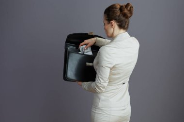 Orta yaşlı bir iş kadınının arkasında evrak çantası ve dolarlarla gri arka planda para paketleriyle görülmüş..