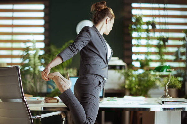 Sustainable workplace. Seen from behind modern business woman in a grey business suit at work stretching leg.