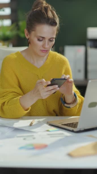 現代緑のオフィスでスマートフォンを使用してラップトップと黄色いセーターの幸せなエレガントな中小企業のオーナーの女性 — ストック動画