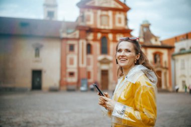Prag Çek Cumhuriyeti 'nde sarı bluz ve yağmurluk giyen şık, yalnız bir turist kadın akıllı telefon kullanıyor ve yürüyor..