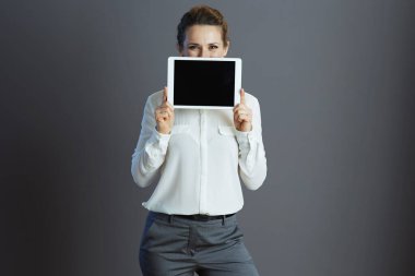 Kırk yaşında beyaz bluzlu genç bir kadın tablet bilgisayar ekranını gri arka planda gösteriyor..