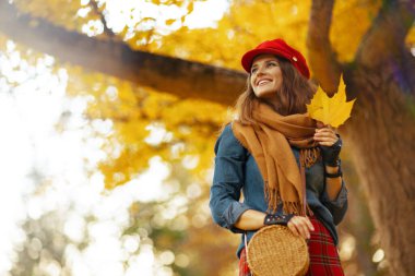 Merhaba sonbahar. Gülümseyen moda kadın kot gömlekli ve kırmızı şapkalı sonbahar yapraklı, eşarplı, eldivenli ve çantalı şehir parkında.