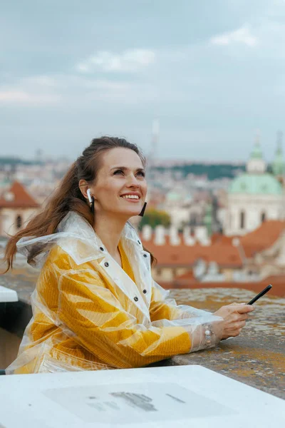 捷克共和国布拉格 身穿黄色衬衫和雨衣 头戴智能手机和耳机 对着城市全景微笑的年轻旅行者 — 图库照片