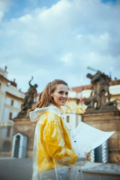 布拉格城堡附近 一名身穿黄色上衣和雨衣的年轻女子带着地图笑着 — 图库照片