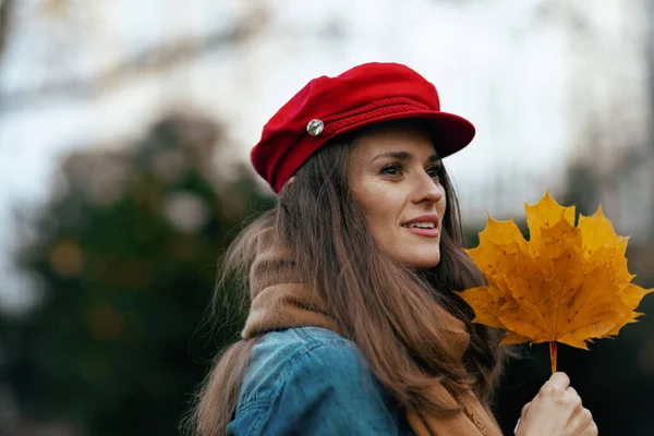 城市里穿着牛仔裤衬衫 头戴红帽子 头戴秋天叶子和围巾的年轻女子 — 图库照片