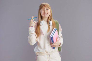 Gri üzerine izole edilmiş sırt çantası, çalışma kitabı, kulaklık ve kalemle bej eşofmanlı mutlu modern kız portresi.