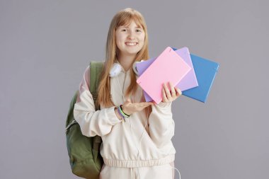 Gri arka planda izole edilmiş sırt çantası, çalışma kitabı ve kulaklığı olan bej eşofmanlı mutlu modern genç kız portresi.