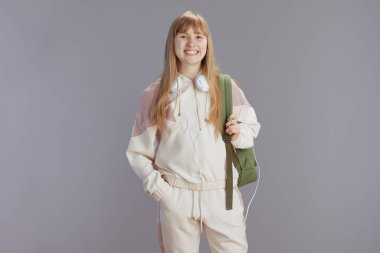 Sırt çantası ve kulaklıkları gri renkte olan bej eşofmanlı, güler yüzlü bir kızın portresi..