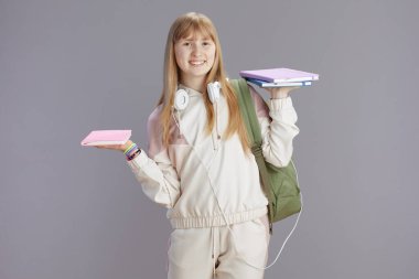 Gri arka planda izole edilmiş sırt çantası, çalışma kitabı ve kulaklığı olan bej eşofmanlı mutlu, modern bir genç kadının portresi..