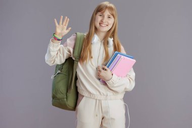 Sırt çantası, çalışma kitabı ve kulaklığı gri arka planda el sallayan bej eşofmanlı mutlu okul kızı portresi.