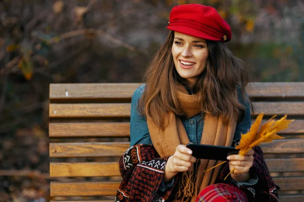 Hello autumn. pensive modern 40 years old woman in red hat with autumn leafs and scarf using smartphone while sitting on bench in the city park.
