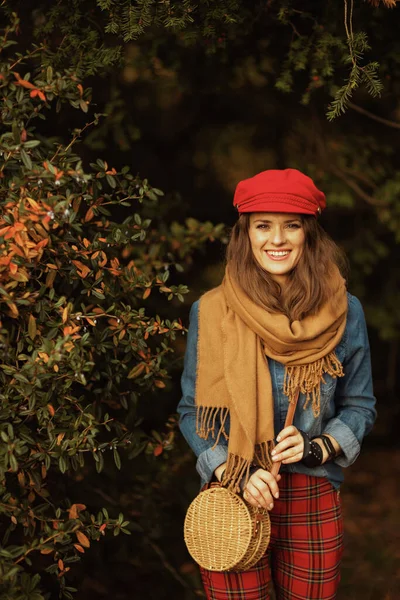 Hello autumn. smiling modern woman in jeans shirt and red hat with scarf, gloves and bag in the city park.