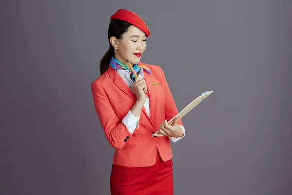 ペンシブ スタイリッシュなアジアの女性のスチュワーデス 赤いスカート ジャケットと灰色の背景に対するクリップボードとの帽子の制服 — ストック写真