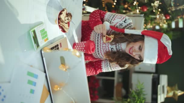 クリスマス タイム サンタの帽子の幸せなエレガントなビジネス女性と伝統的なクッキーとラップトップとクリスマスツリーと現代緑のオフィスでバーチャルミーティングを持つ赤のクリスマスセーター — ストック動画