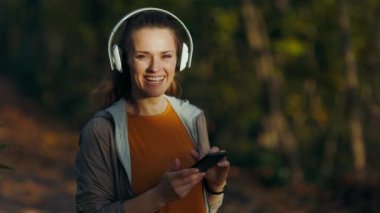 Merhaba sonbahar. Gülümseyen şık orta yaşlı kadın parkta spor kıyafetleri içinde kulaklıkla müzik dinliyor..