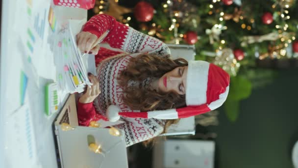 クリスマス タイム クリスマスツリーと現代グリーンオフィスで働く文書とラップトップとサンタの帽子と赤のクリスマスのセーターで現代の小さなビジネスオーナーの女性を強調 — ストック動画