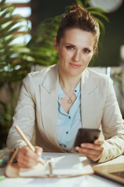 Modern yeşil ofisteki hafif bir iş elbisesi içindeki modern kadın çalışanın portresi. Akıllı telefon uygulamaları kullanarak defter..