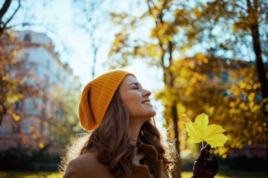 Merhaba Eylül. Mutlu, orta yaşlı, kahverengi ceketli, sarı şapkalı, güz sarısı yapraklı bir kadın sonbaharda şehir sokaklarında..