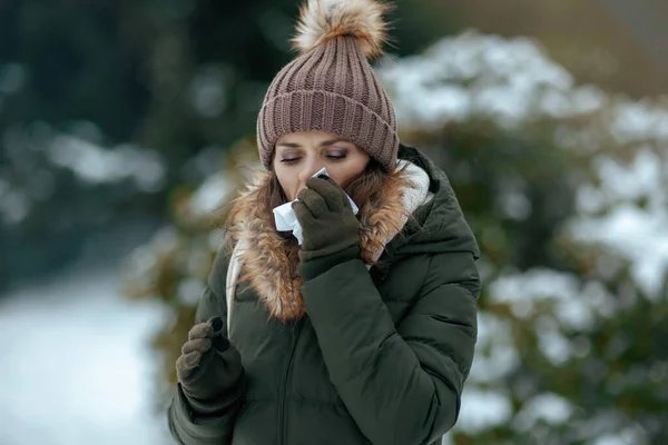 sad modern 40 years old woman in green coat and brown hat outdoors in the city park in winter with mittens, napkin blowing nose and beanie hat.