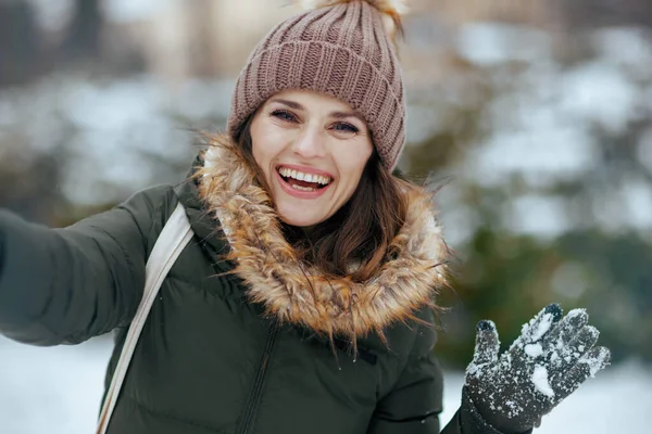 happy modern woman in green coat and brown hat outdoors in the city park in winter with snowy mittens and beanie hat having online meeting.