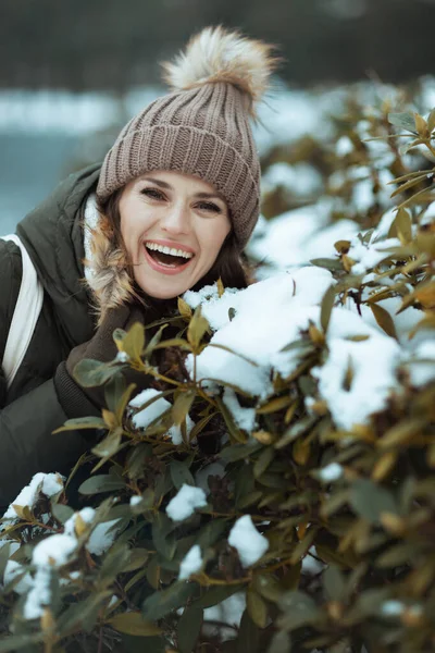 雪の枝の近くのビーニー帽子と冬の公園の緑のコートと茶色の帽子の現代中年の女性を微笑みます — ストック写真