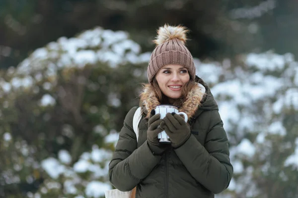 快乐的现代女性 冬天穿着绿色外套 头戴褐色帽子 头戴手套 拿着豆奶拿铁和豆帽 站在雪地的枝头附近 — 图库照片
