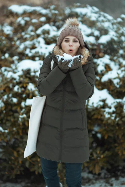 modern woman in green coat and brown hat outdoors in the city park in winter with mittens and beanie hat near snowy branches.