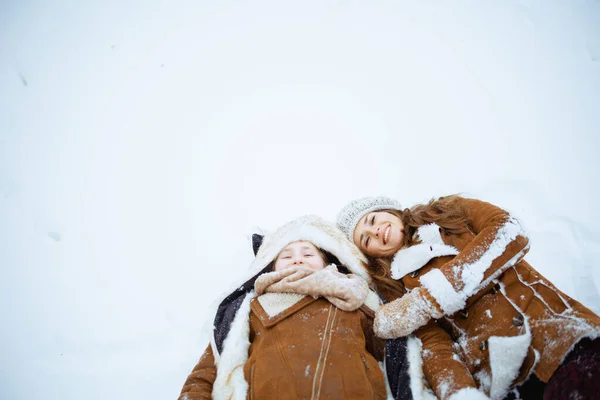 happy modern mother and daughter in coat, hat, scarf and mittens laying in snow outdoors in the city park in winter.