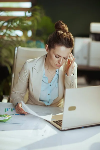 stressed modern woman worker in a light business suit in modern green office with documents and laptop.
