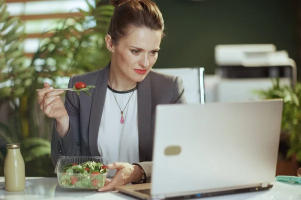 Sustainable workplace. modern 40 years old small business owner woman in a grey business suit in modern green office with laptop eating salad.