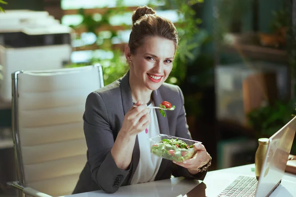 持続可能な職場 幸せな現代中年の女性の労働者は現代緑のオフィスの灰色のビジネス スーツでサラダを食べるラップトップと — ストック写真