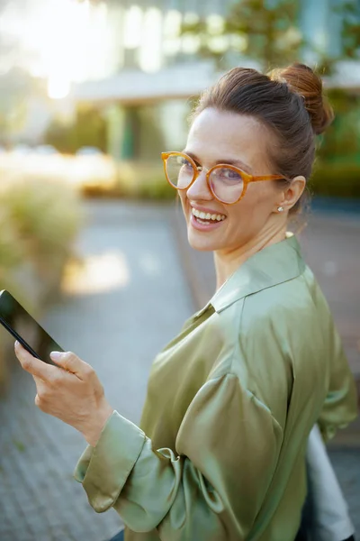 在商业区穿着绿色衬衫和眼镜的现代女性员工用智能手机微笑 — 图库照片