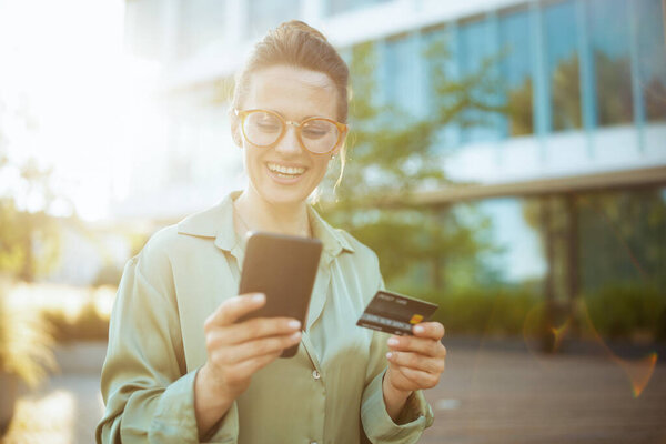 smiling modern woman worker in business district in green blouse and eyeglasses with credit card using smartphone.