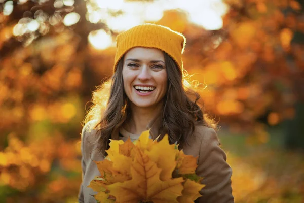 Hello autumn. smiling trendy middle aged woman in brown coat and yellow hat with autumn yellow leaves outside in the city park in autumn.