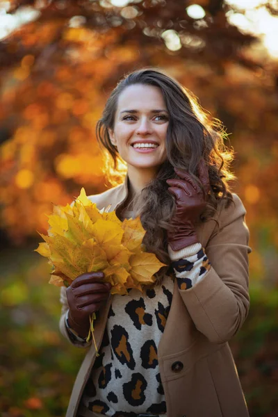 Hello october. smiling trendy 40 years old woman in brown coat with autumn yellow leaves outdoors in the city park in autumn.