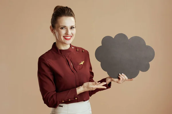 happy stylish female air hostess against beige background with cloud shaped blackboard.