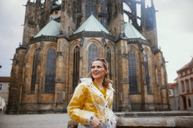 Prag Çek Cumhuriyeti 'nde sarı bluzlu ve yağmurluklu mutlu modern yalnız turist kadın yürüyüş turunda..