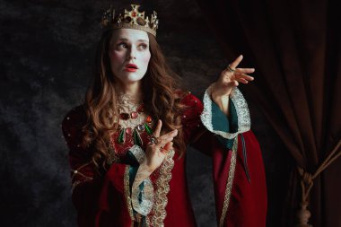 Kırmızı elbiseli, beyaz makyajlı ve tacı koyu gri arka planda olan ortaçağ kraliçesi..